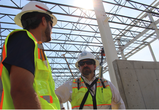 Two IES employees looking at each other in safety gear standing below steel crossbeams
