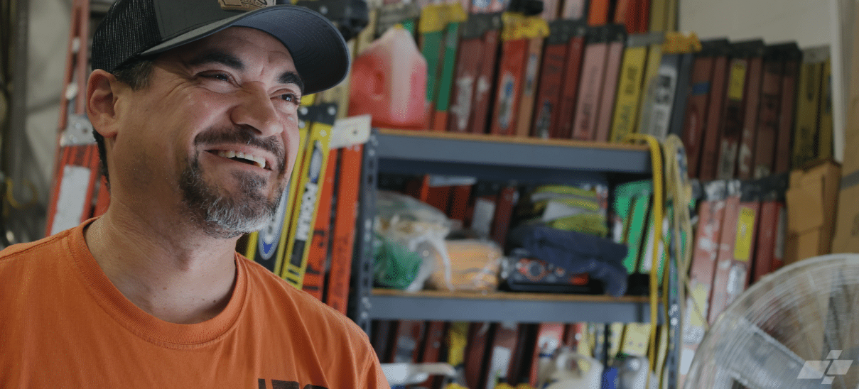 Man in orange shirt and hat smiling off camera with ladders behind him
