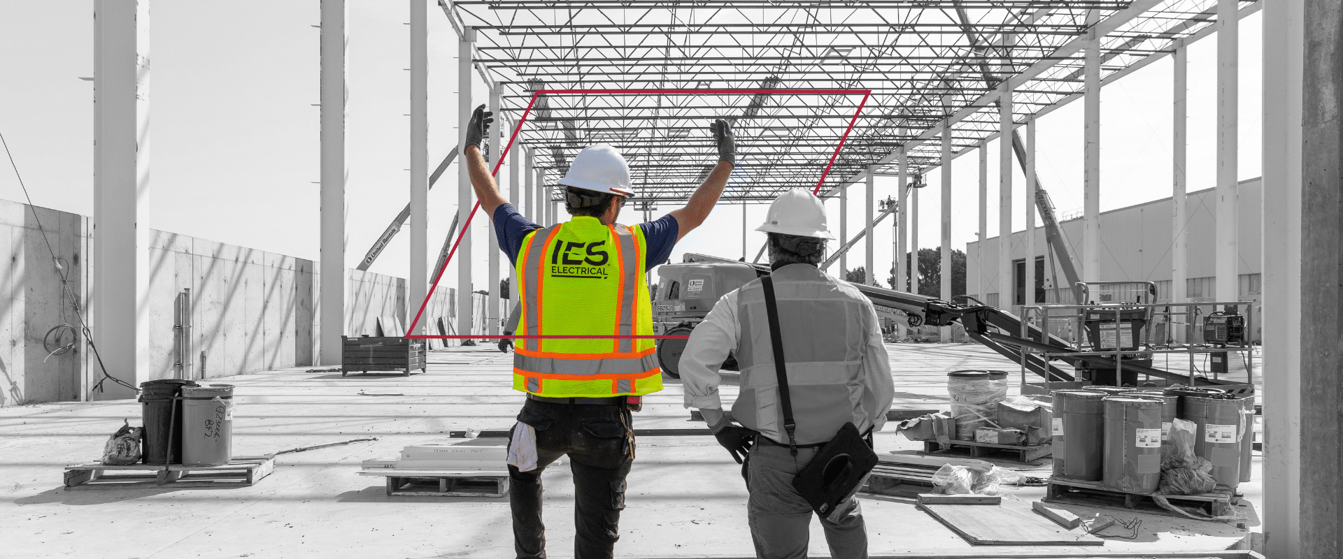 Black and white image of steel framing construction site with two IES employees - one in full color and safety gear