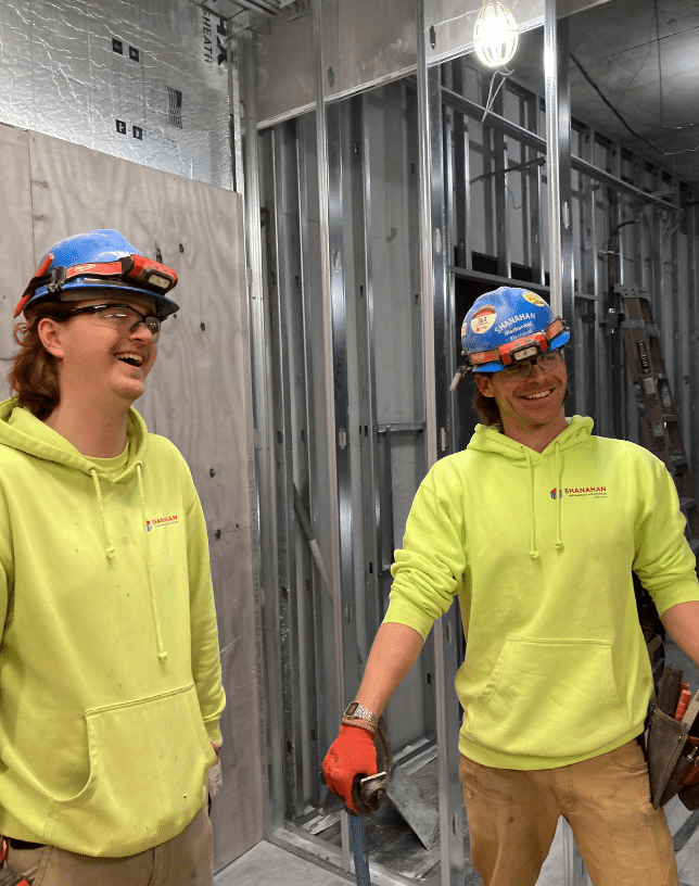 Two men in yellow safety sweatshirts and blue hardhats laughing