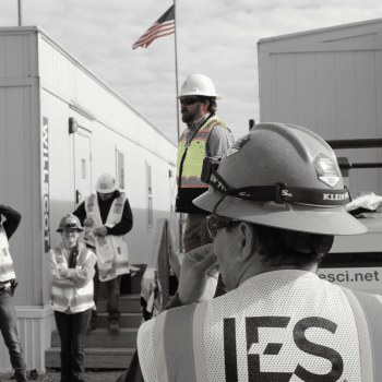 Black and white image of IES employees listening to man on pedestal in front of american flag