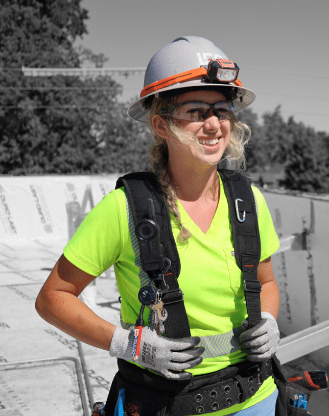 Woman in yellow shirt and harness and hardhat smiling at jobsite